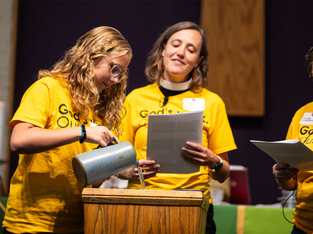 A teenage female with blonde hair pours water from a clay pitcher into a square baptismal font as a woman in pastor’s collar stands behind her and reads from a paper bulletin. 