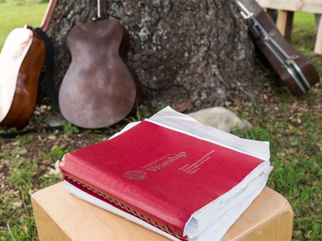 A person in a yellow T-shirt, white headscarf and green dress carries a black bucket in their left arm as they walk away from the camera, next to another person in a yellow shirt and blue pants carrying an aqua bucket in their left hand.