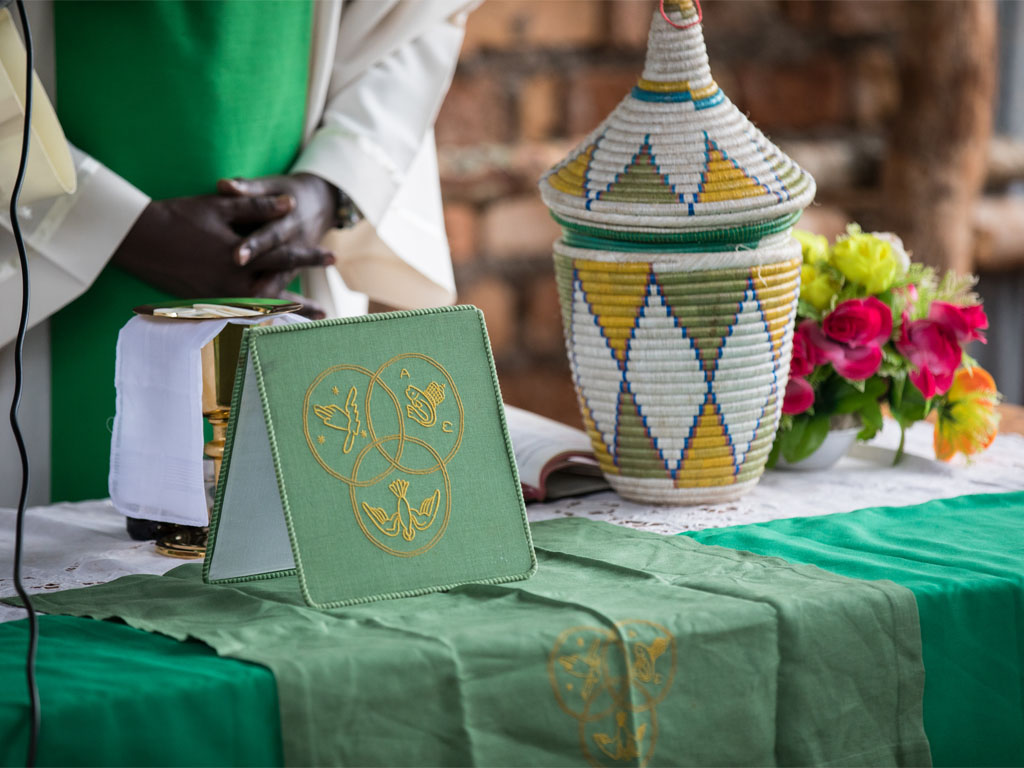 Person in a white robe with green pastoral vestments stands with hands folded behind an altar with a woven jar.