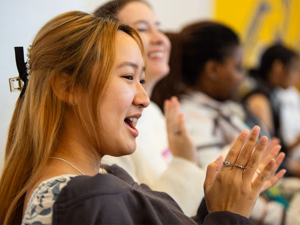 A woman with long red-brown hair claps as she sits in a row with others out-of-focus. 