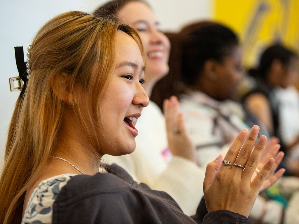 A woman with long red-brown hair claps as she sits in a row with others out-of-focus. 