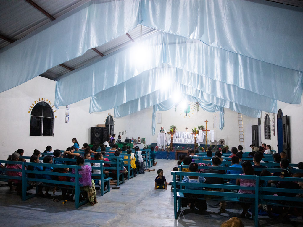 people sit on blue benches looking toward a white-draped altar in a white adobe room with white curtains hanging from the ceiling.