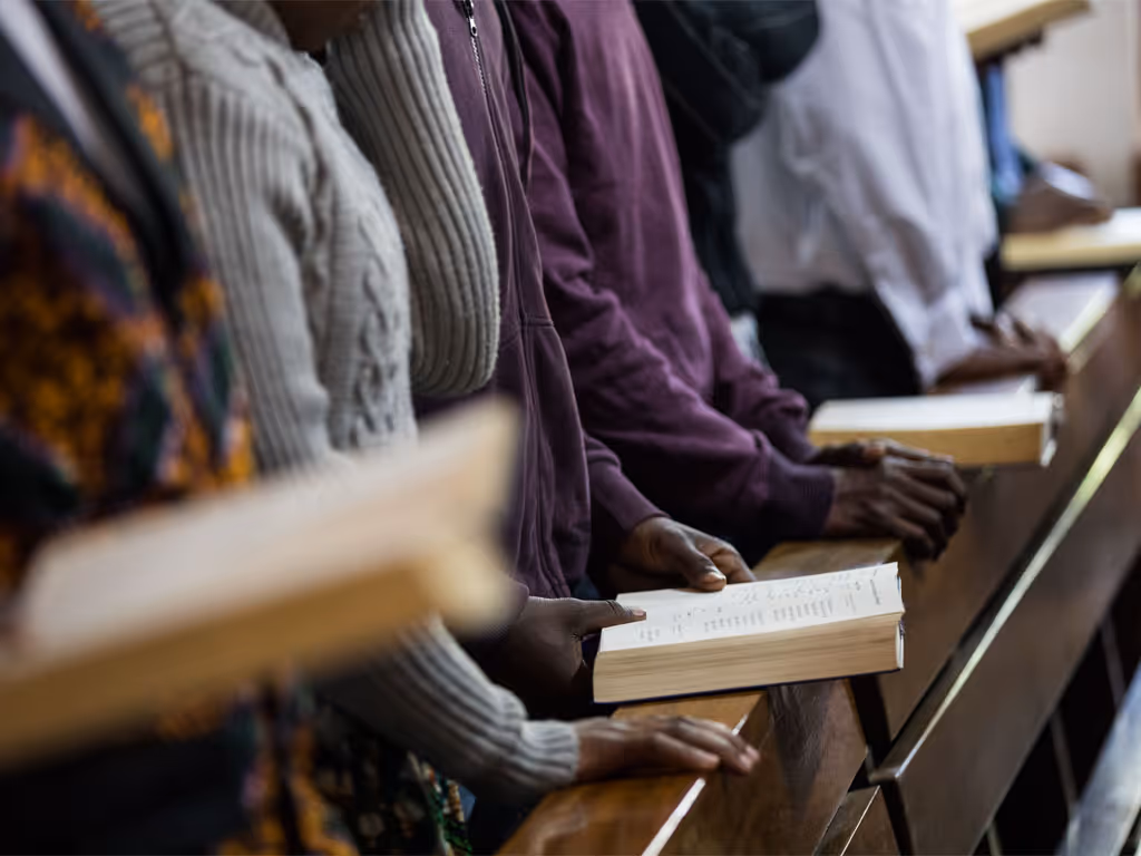 a row of people, faces not shown on camera, stand leaning against a wooden railing with hymn books open and hands folded in prayer. 