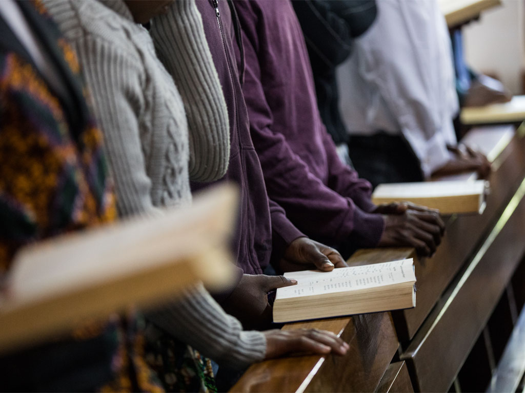 a row of people, faces not shown on camera, stand leaning against a wooden railing with hymn books open and hands folded in prayer. 
