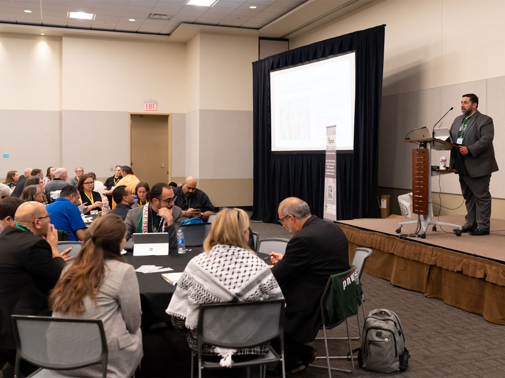 A man in a gray suit stands at a podium looking out at a group of people gathered at round tables with black tablecloths.