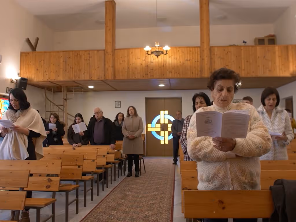 Small group of people on either side of a carpeted aisleway holding paper bulletins as they sing songs in a church with a wooden balcony behind them.