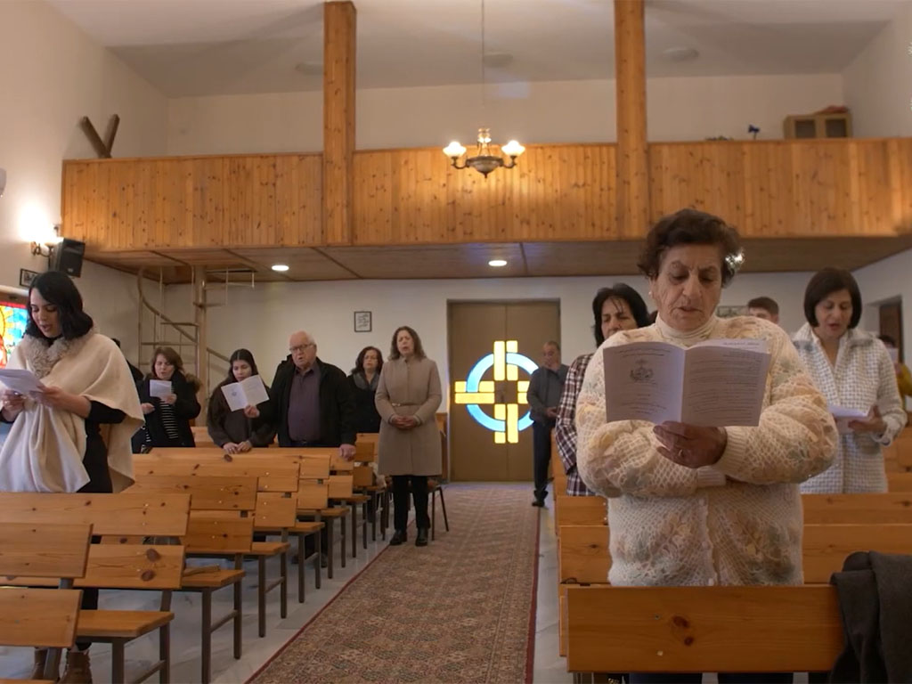 Small group of people on either side of a carpeted aisleway holding paper bulletins as they sing songs in a church with a wooden balcony behind them.
