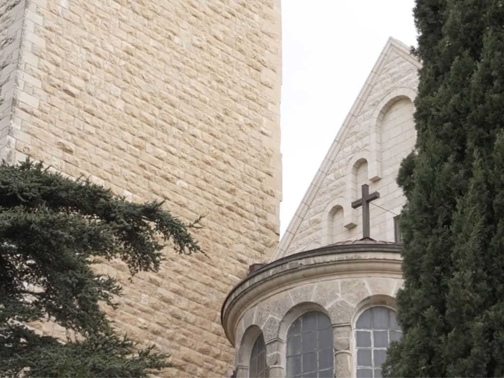 Stone pillar next to a rounded roof with a cross on top with green trees on either side. 