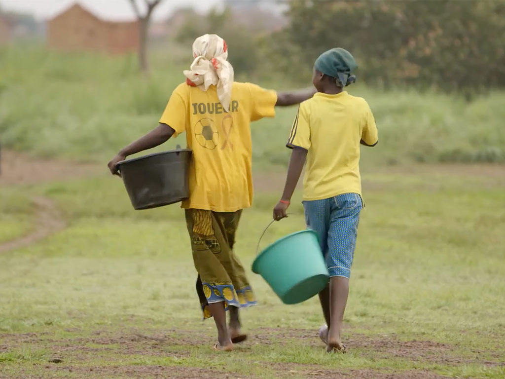 A person in a yellow shirt with white headscarf and green dress carries a bucket in their left arm as they walk with another person in a yellow shirt, blue pants and green hair wrap carrying a green bucket in their left hand. 