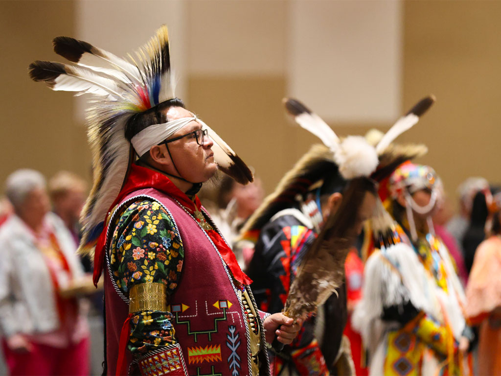 A man wearing a Native American headdress holds a large feather as he stands in a line with other, similarly dressed people. 