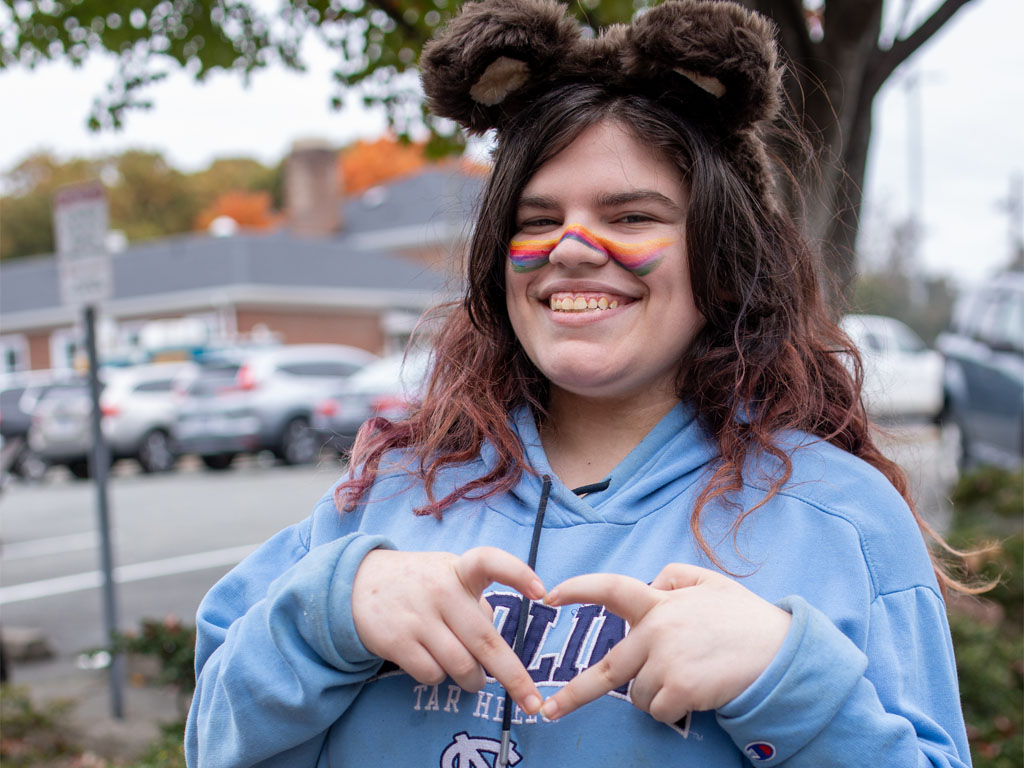 Person in a blue sweartshirt with striped facepaint and furry ear headband makes a hart with their hands.  
