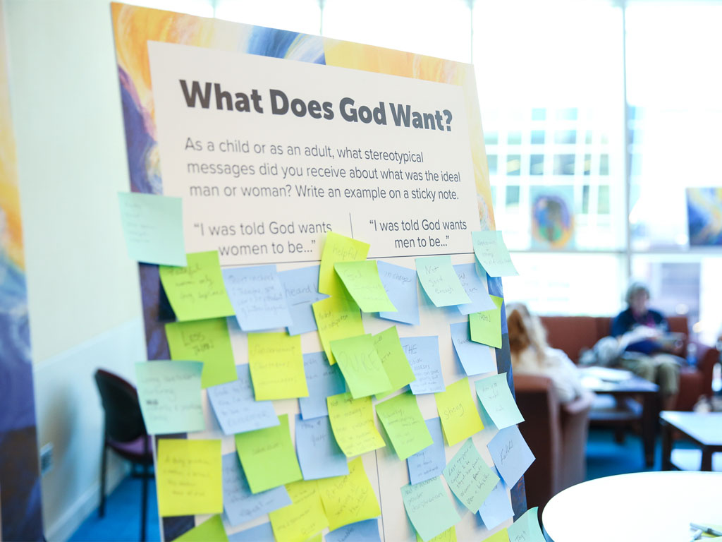 A poster board with the title “What Does God Want?” and subheadings “I was told God wants women to be” and “I was told God wants men to be” is filled with post-it notes in yellow, green and blue.