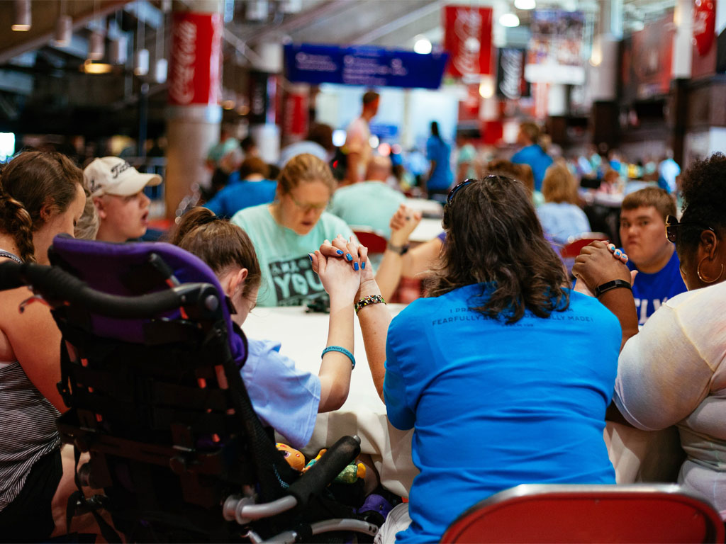 Group of teenagers sitting around a circular table holding hands in prayer; a female with her back to the camera is sitting in a high-backed wheelchair.  