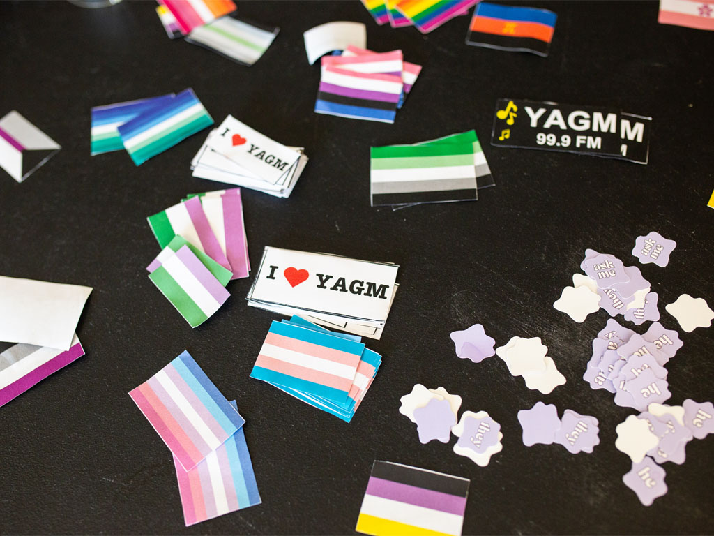 Stickers of various pride flags, purple starbursts with pronouns, and white stickers with “I heart YAGM” sit on a black table.