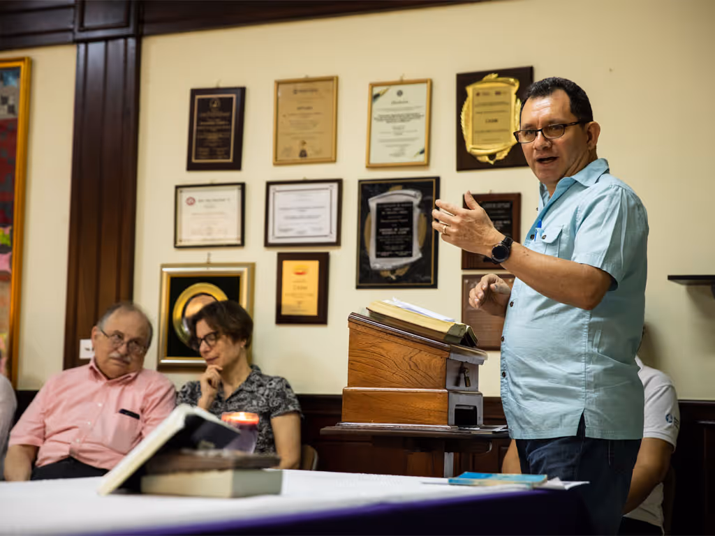 A man with short black hair and glasses wearing a light blue polo shirt gestures with his left hand as he reads from a bible on a podium in front of him. 