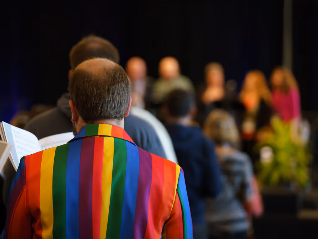 Male figure looking away from the camera wears a collared shirt with vertical rainbow pattern while holding a hymnal during a worship service.  