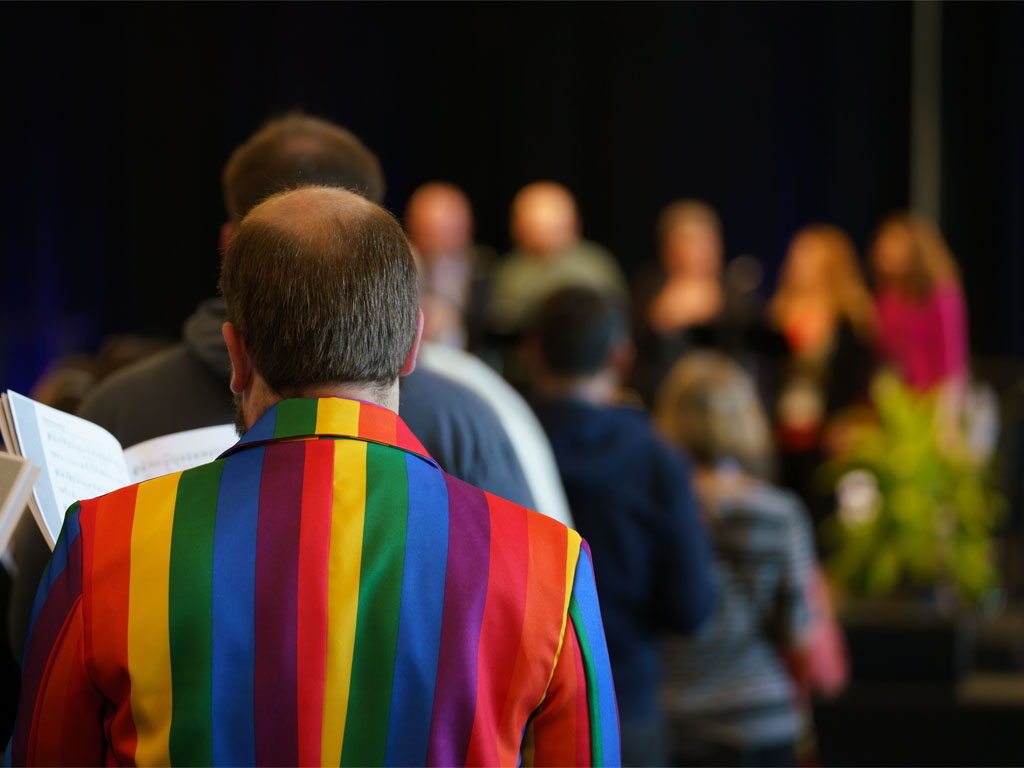 Male figure looking away from the camera wears a collared shirt with vertical rainbow pattern while holding a hymnal during a worship service.  