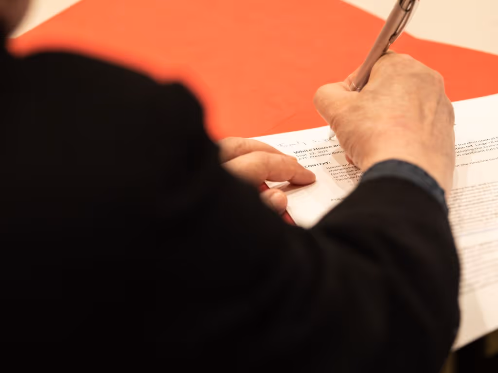 An older man with white hair and a mustache, wearing a yellow shirt that reads “God’s work. Our hands.” in black text, shakes hands with a young female as she walks out of a church sanctuary.
