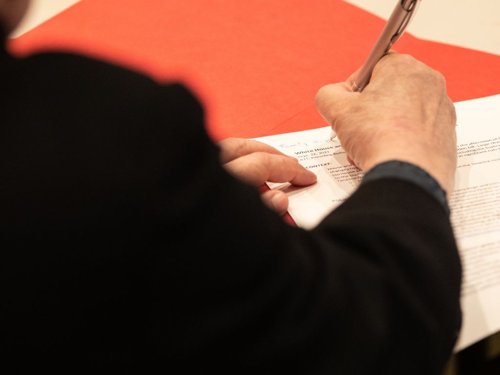 An older man with white hair and a mustache, wearing a yellow shirt that reads “God’s work. Our hands.” in black text, shakes hands with a young female as she walks out of a church sanctuary.