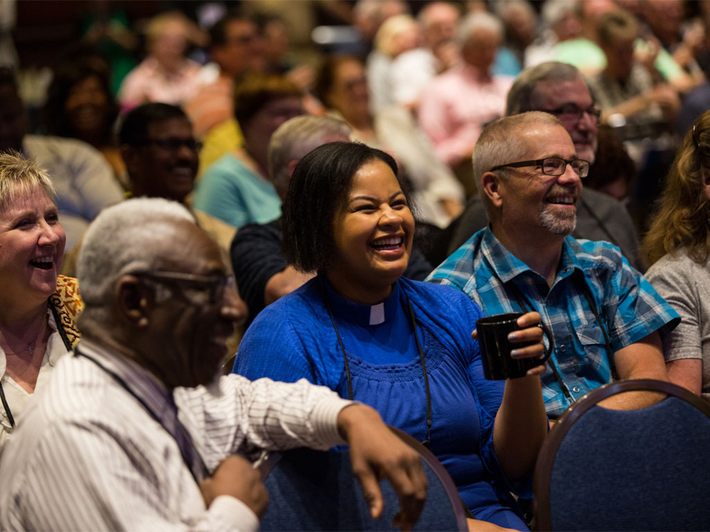 A woman in a blue top with a white pastoral collar laughs as she looks at a stage along with a man in a blue plaid shirt, a man in a white striped dress shirt, and a woman in a white shirt with yellow patterened overshirt. 