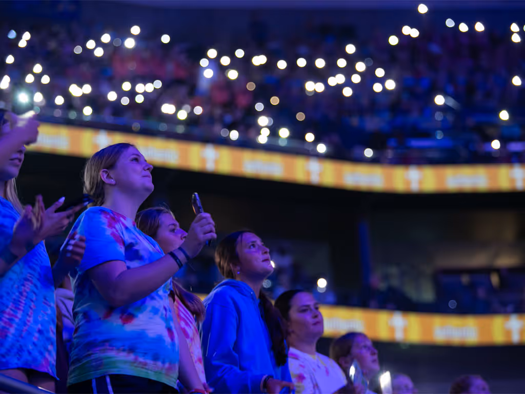 A row of teenage girls hold their phones in the air inside an arena lit in dark blue with cell phone lights blinking around them. 