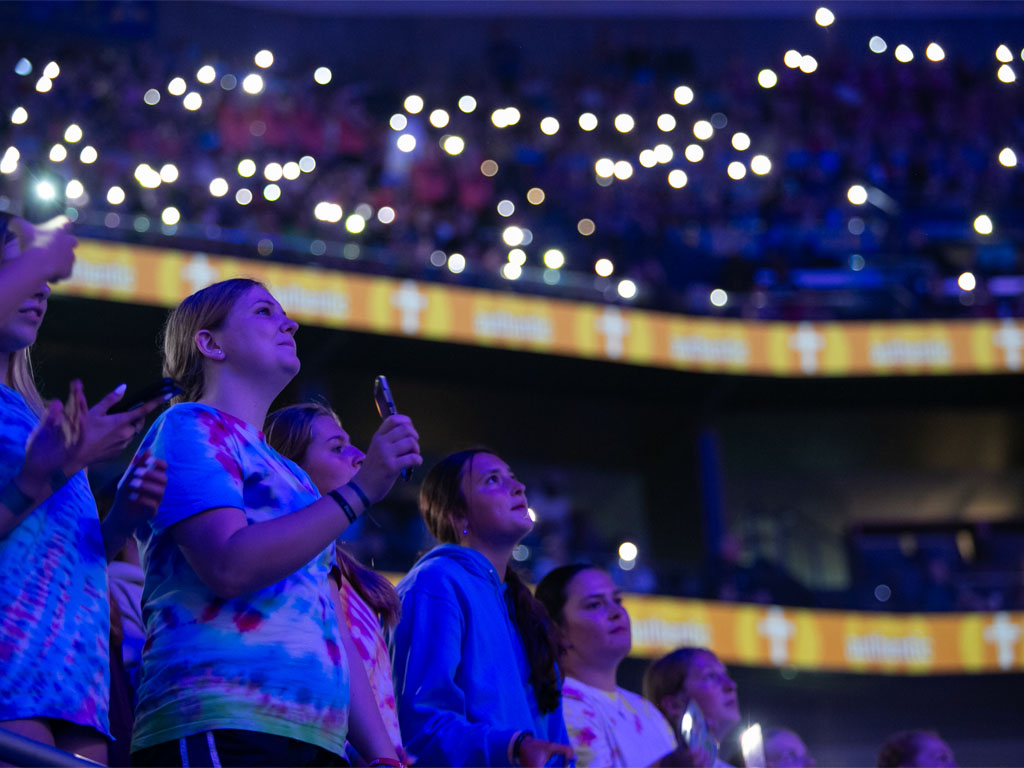 A row of teenage girls hold their phones in the air inside an arena lit in dark blue with cell phone lights blinking around them. 