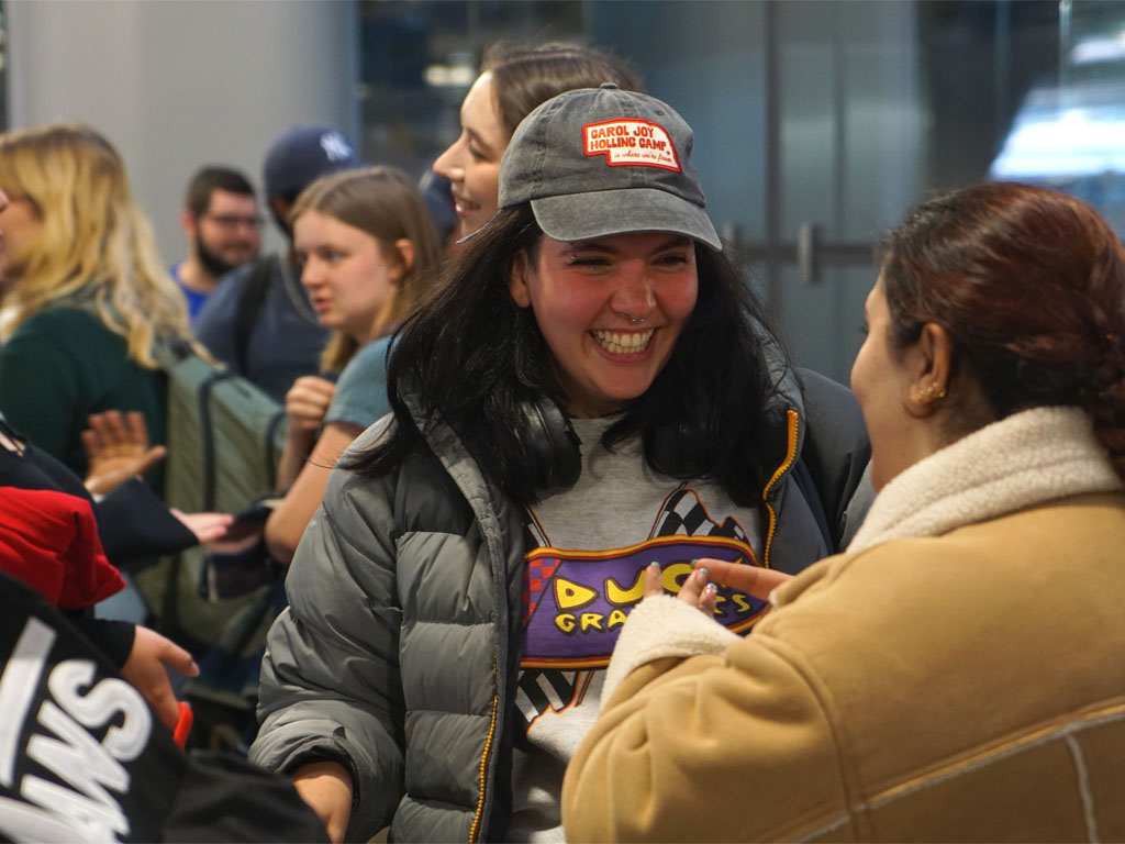 College-age female with a puffy gray jacket and gray baseball cap talks to another person whose back is to the camera wearing a sheepskin-like coat in a crowded room with other young adults. 