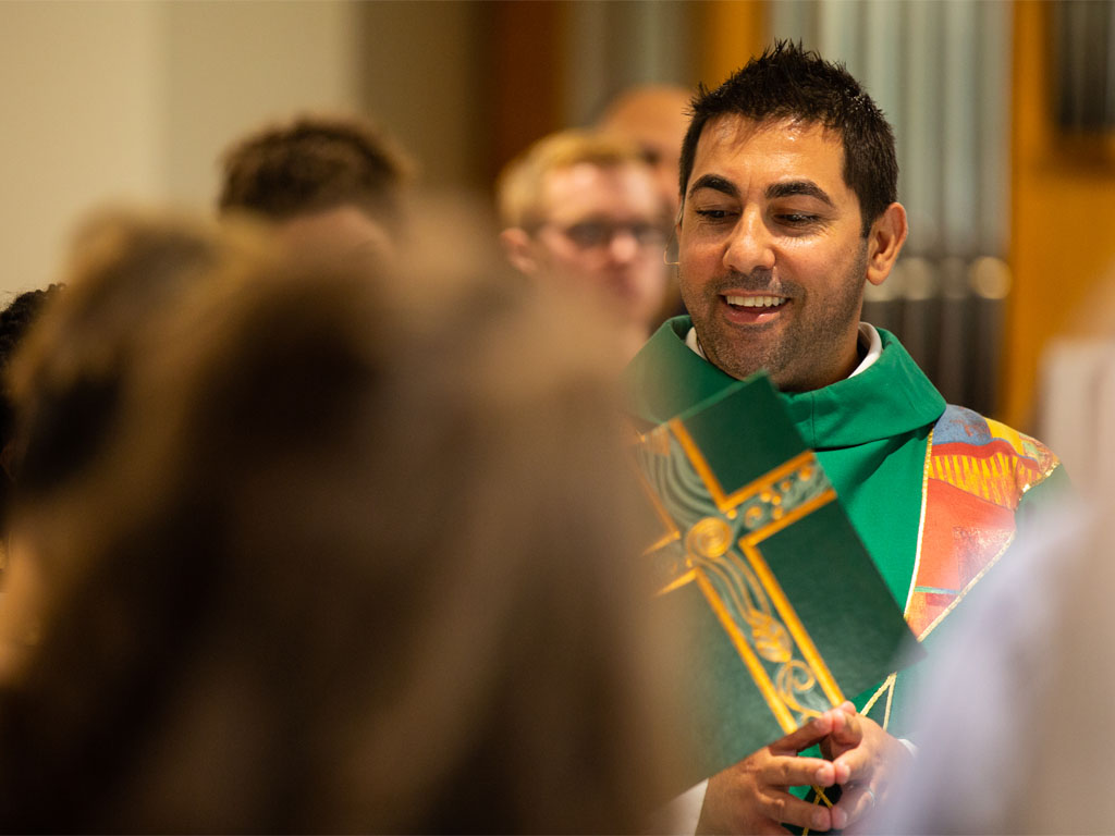  A man with short black hair wearing a white robe and pastor’s vestments reads from an open green book with yellow cross among a group of out-of-focus people during a worship service.