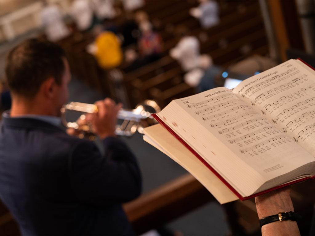 red worship book open to two hymns in front of man playing a trumpet as the image looks out to an out-of-focus congregation in church pews. 