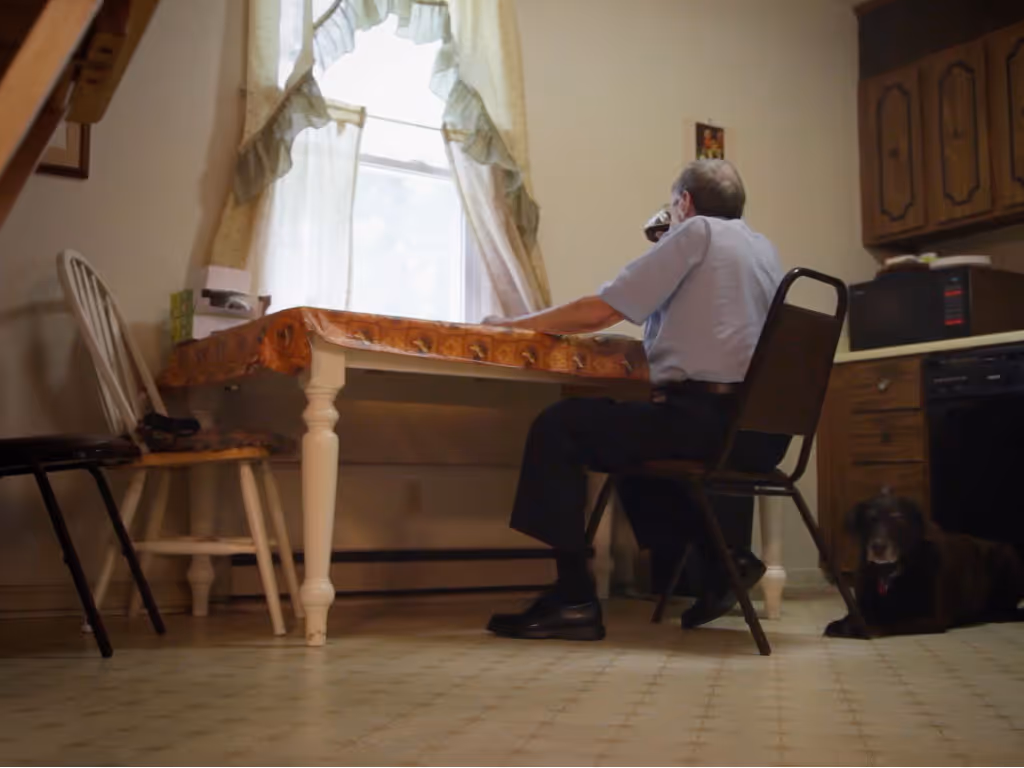 A man sits alone at a table looking out a window while drinking a can of soda.
