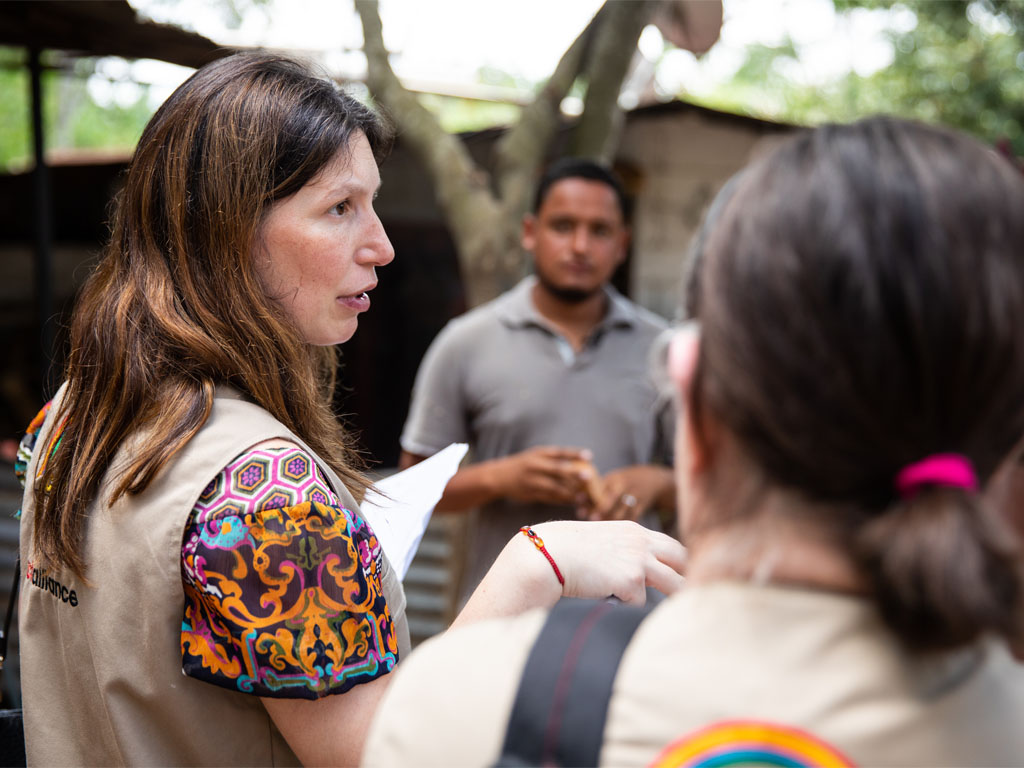 Woman wearing a tan vest over a colorful shirt speaks with a group of people gathered in a circle outdoors in front of a tree and a small hut. 