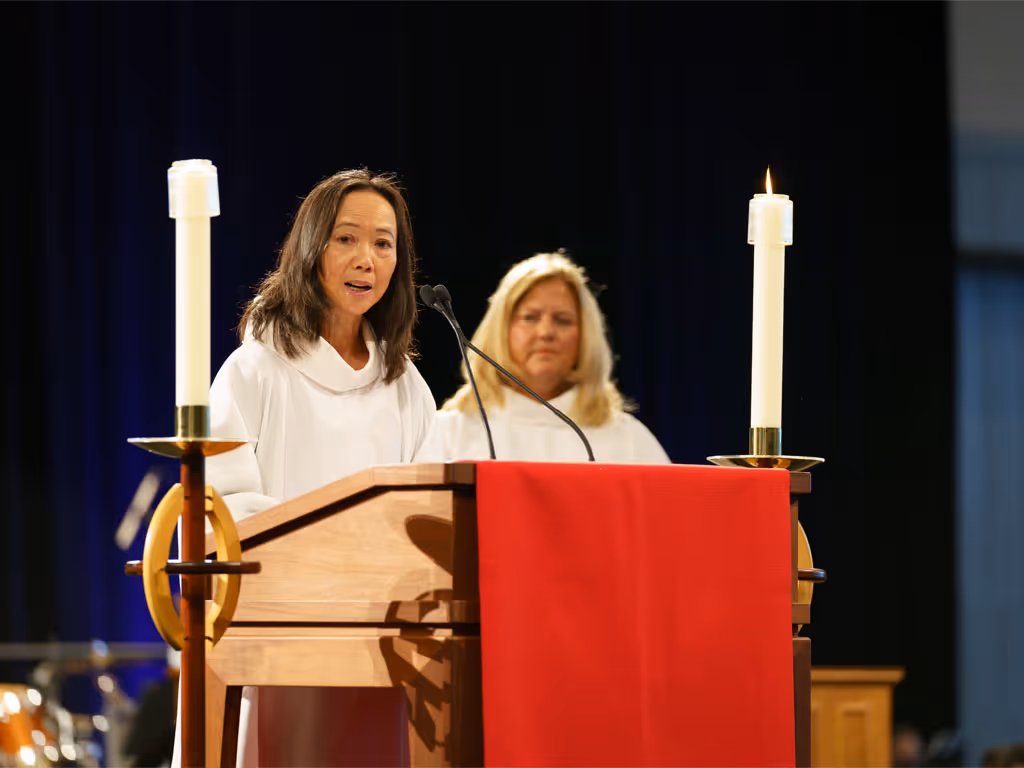 Woman with long dark hair in white robe preaches from a podium with red parament with candles on either side as another woman with blonde hair stands in the background.