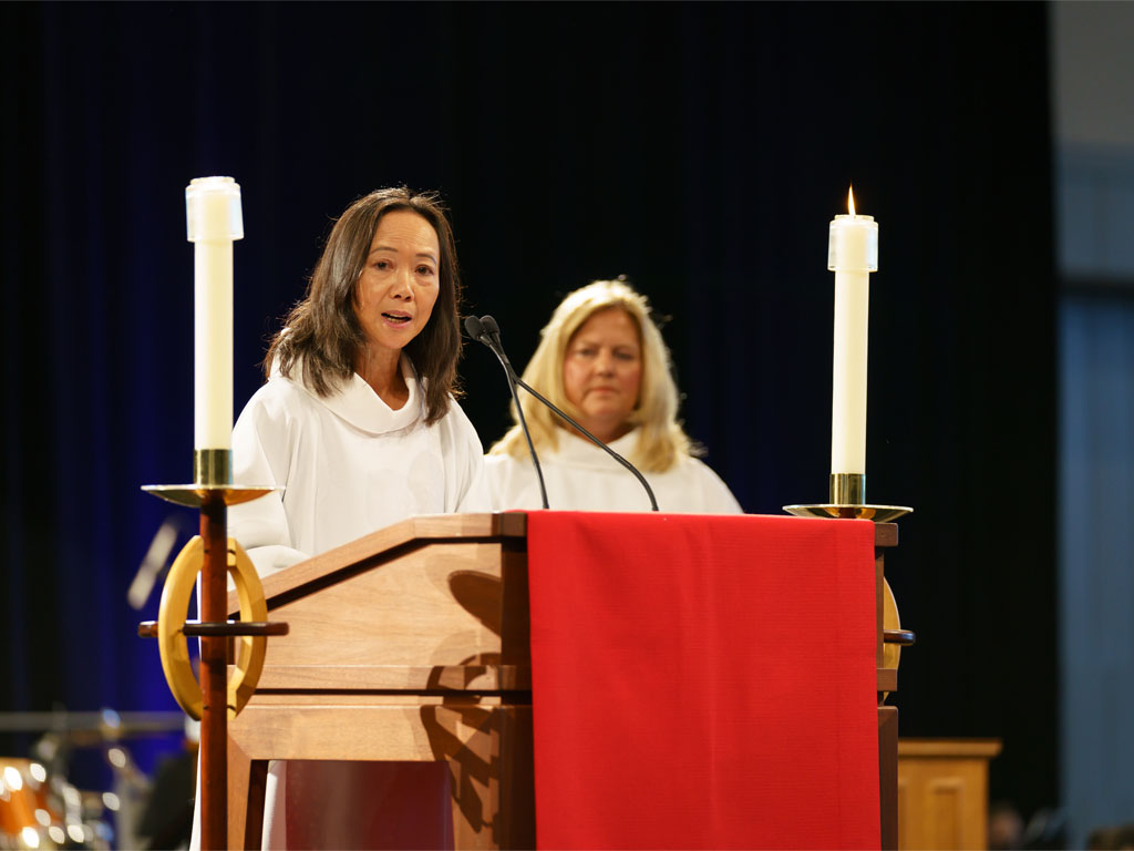 Woman with long dark hair in white robe preaches from a podium with red parament with candles on either side as another woman with blonde hair stands in the background.