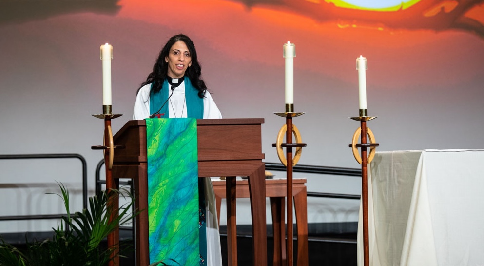 Woman with long black hair wearing a white robe with green vestments preaches at a podium with a candle to the left and two candles to the right.