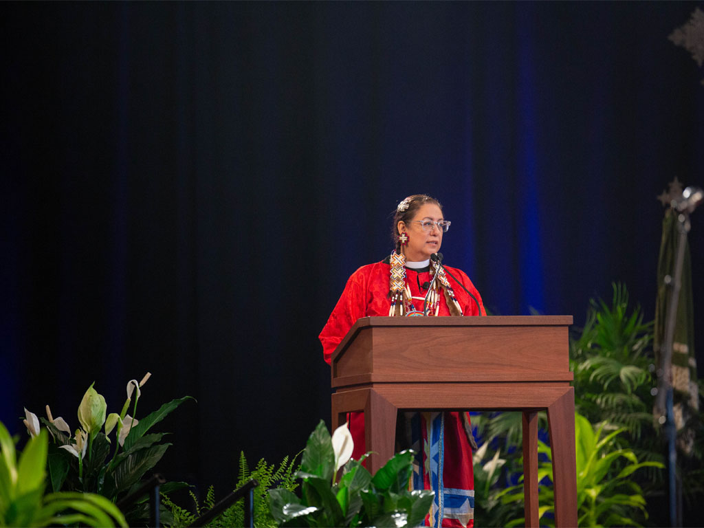 Woman wearing a red Native American dress with beaded vestment around her neck stands at a podium in front of a blue wall with green plants all around.