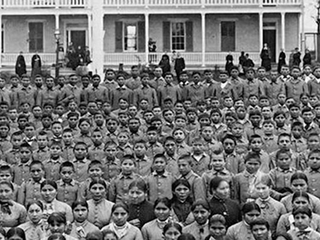 black and white photograph of rows of indigenous youth in school uniforms standing in front of a Victorian-style building.