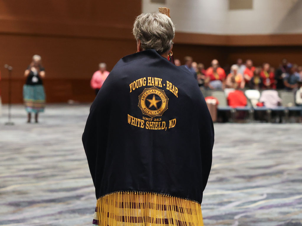 Single figure wearing a black jacket with gold letters and gold fringe stands facing away from the camera in a large open room. The back of the jacket reads “Young Hawk Bear. White Shield, ND.”