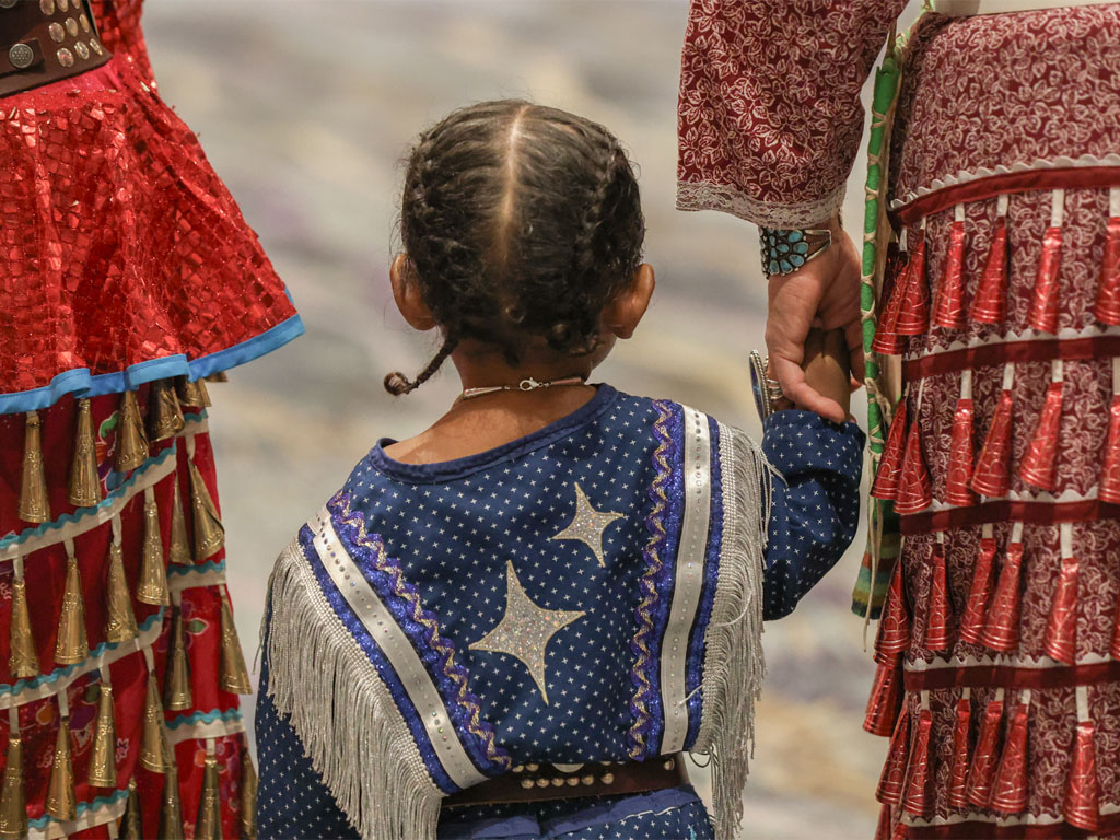 A small child with braided hair wearing a blue Native American dress looks away from the camera while holding the hand of an adult wearing a red dress