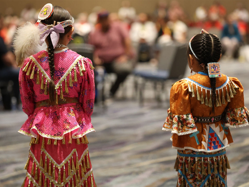 Two young girls in Native American dress – one pink and one gold, with their hair braided look away from the camera toward a room full of out-of-focus people.  Tile 2 image: native_voices_profile_Shannon_Klescewski