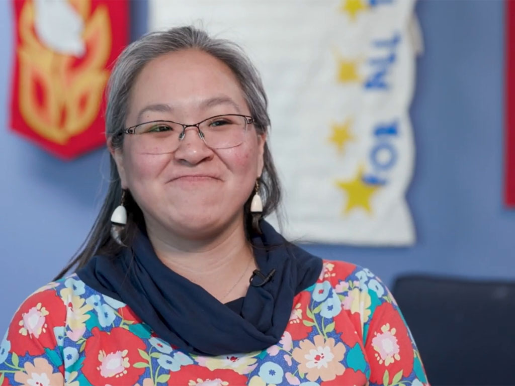 A woman with white hair and glasses wearing a floral print shirt sits in front of a blue wall with a white wall hanging as she smiles closed-mouthed at the camera.