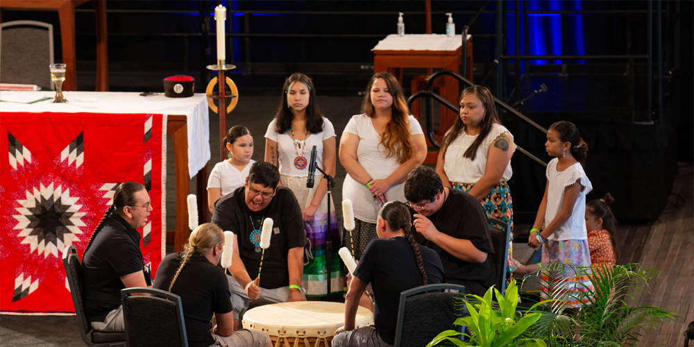 Man in a pink shirt with an open gray sweater and a nametag that reads “Vance” stands behind a glass podium next to a table with blue parament and fabric candles. 