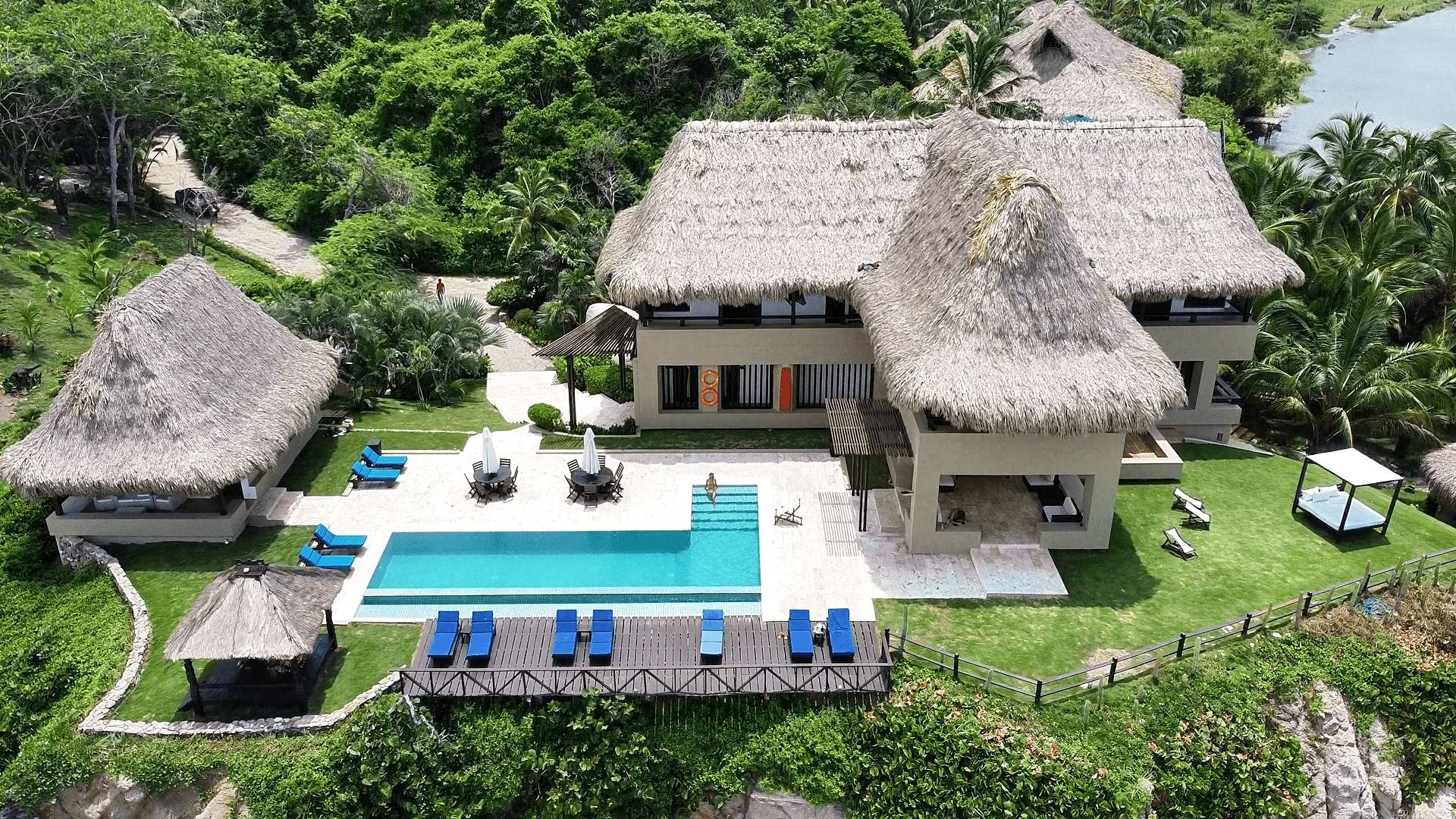 Drone view of a luxury eco-resort in Colombia with a thatched-roof villa, infinity pool, cabanas, and tropical jungle surroundings — perfect for a high-end wellness getaway.