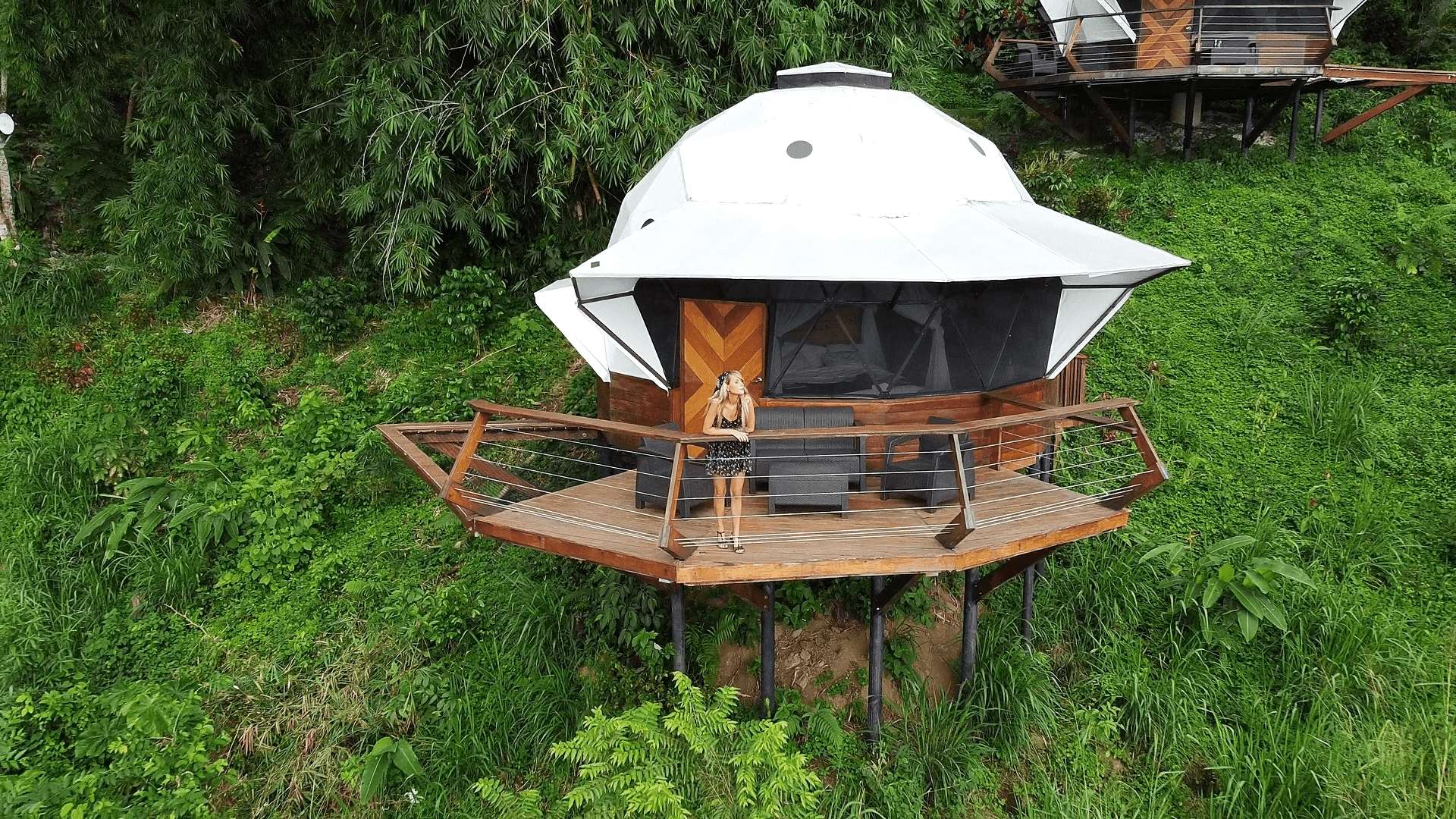  Dome-shaped eco-cabin perched on a lush hillside in Colombia, with a woman standing on the private deck — unique glamping stay perfect for nature and wellness travel.