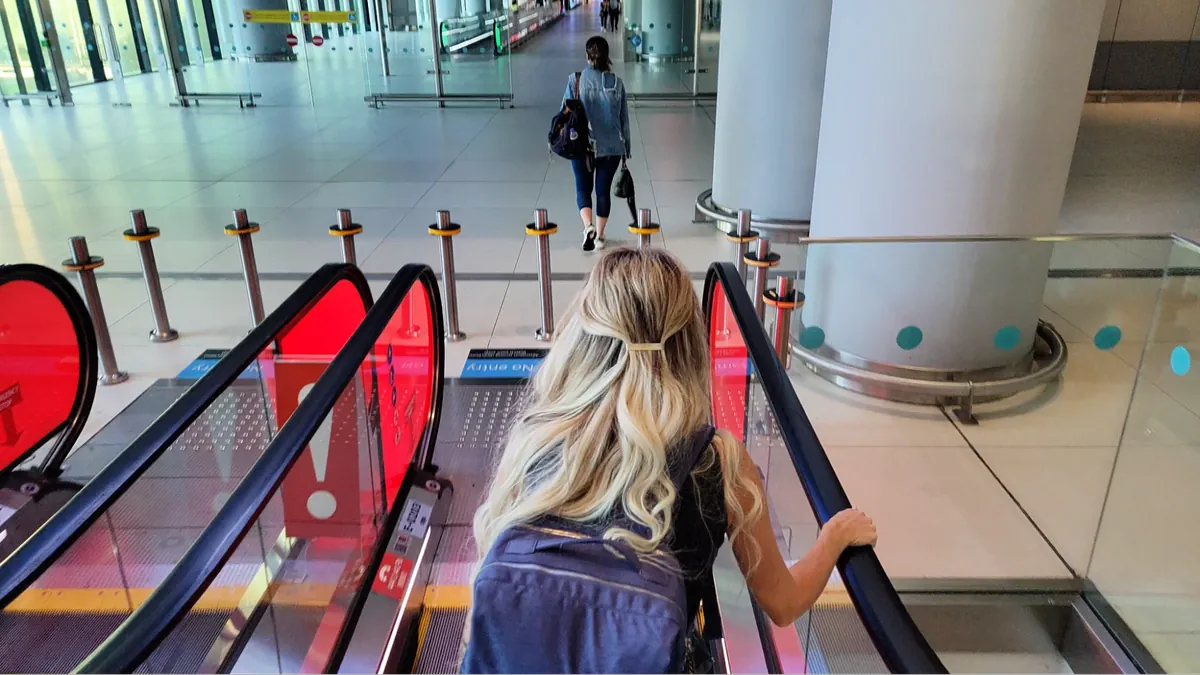 Blonde woman with backpack riding escalator down to entrance area of modern Istanbul Airport terminal.
