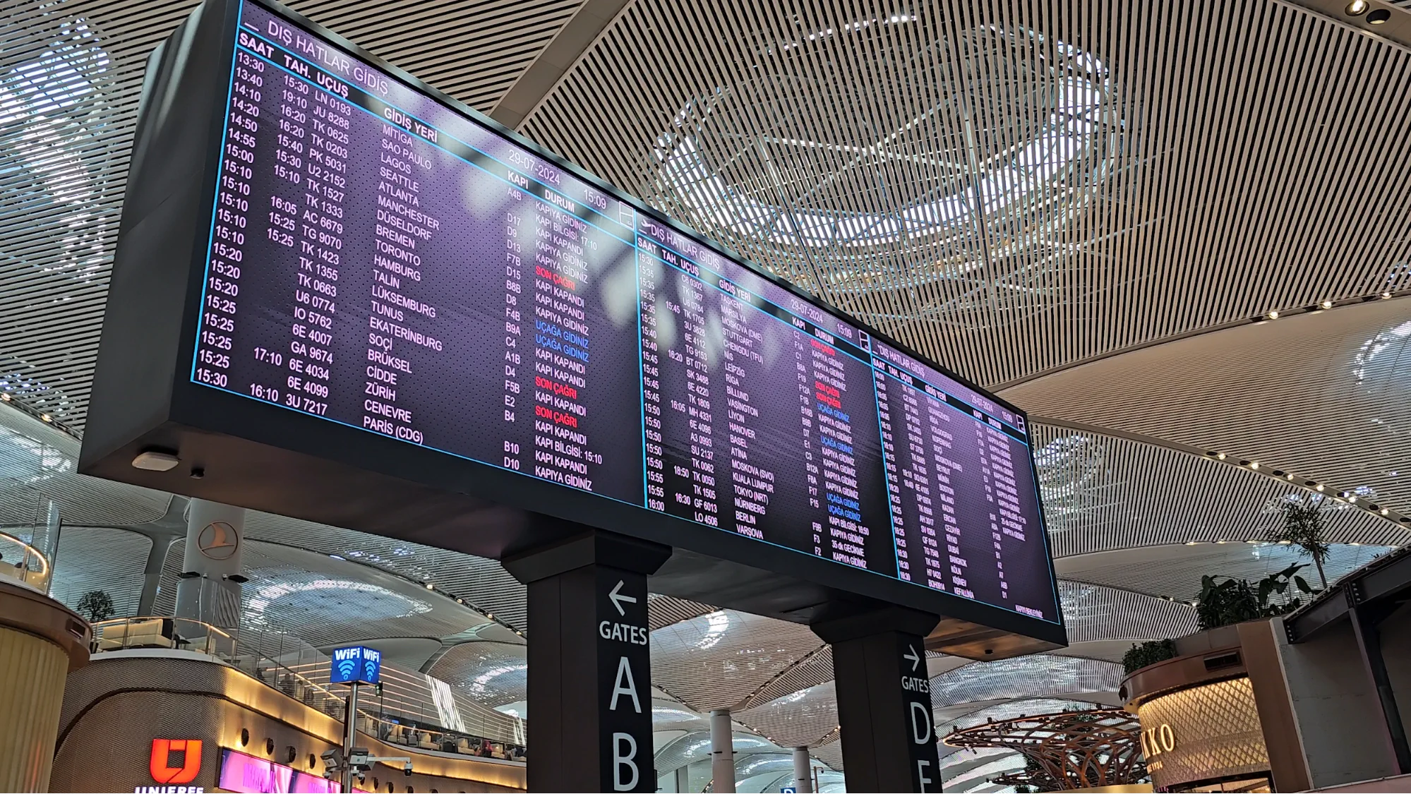 A large departure board inside Istanbul Airport shows international flight times beneath a futuristic ceiling.