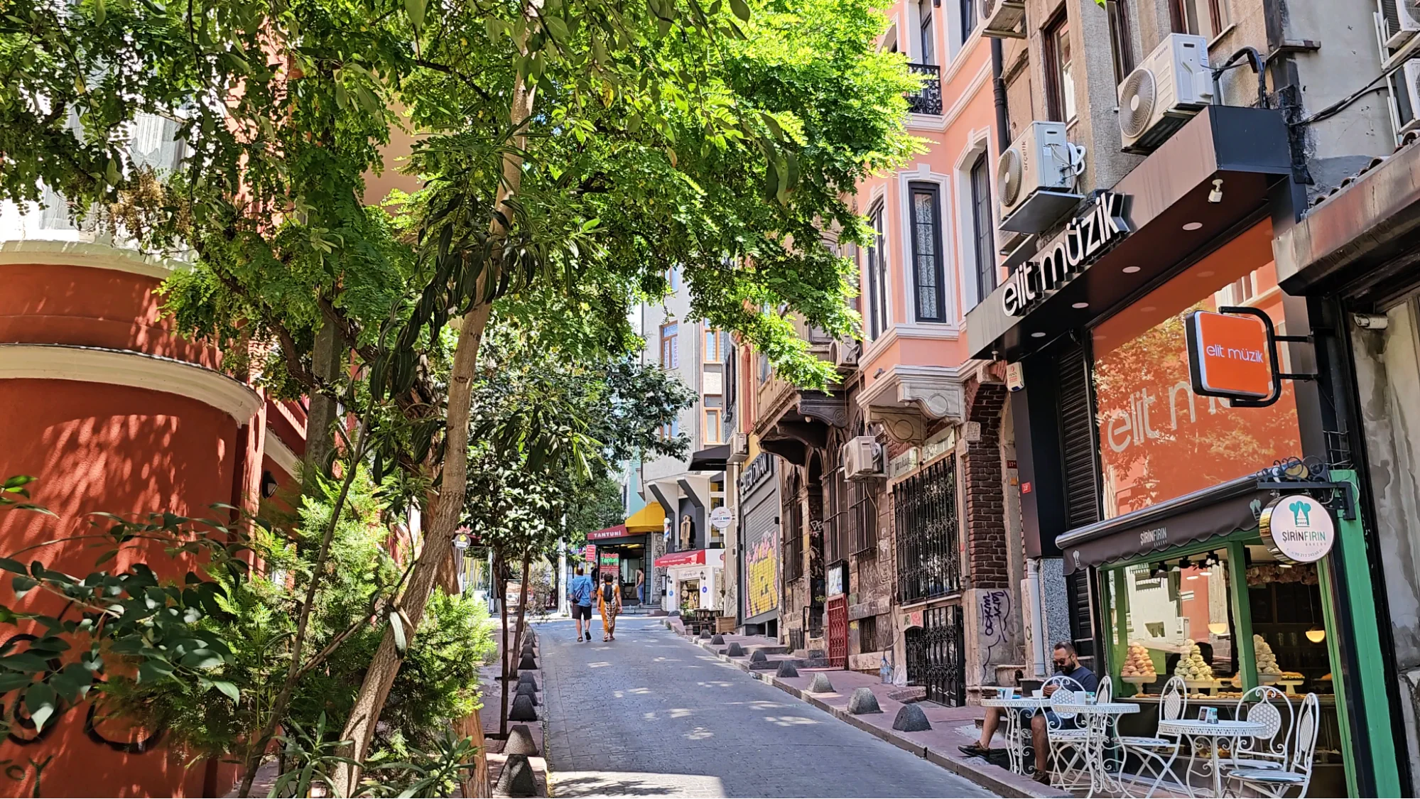 A tree-lined uphill street in Istanbul shows boutique shops, cafes, and pastel-colored buildings with outdoor tables and chairs.