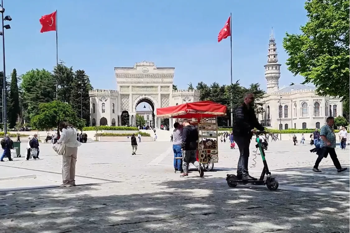 A wide shot of Istanbul University’s grand entrance gate with Turkish flags flying, people walking, and a red cart in the foreground.