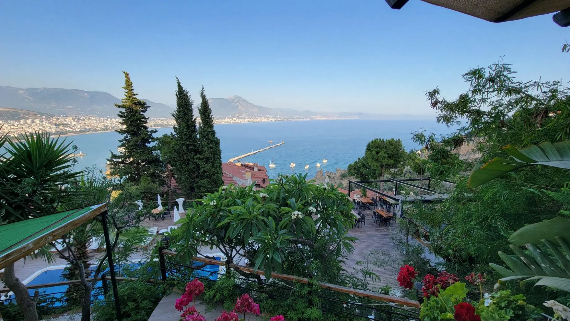 An elevated garden terrace with flowers and lush greenery overlooks Alanya’s coastline, the sea, and surrounding hills and buildings.