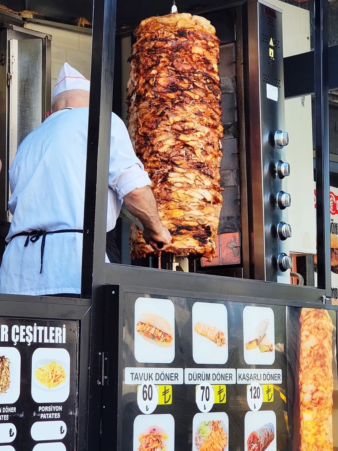 A chef carves meat from a large rotating döner spit at a street stand in Türkiye, with a visible menu of döner options and prices.