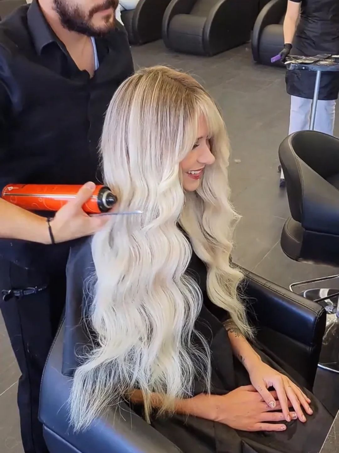 A hairstylist sprays finishing product on long, voluminous, wavy blonde hair of a smiling woman seated in a modern salon chair.	A professional hairstylist perfects glamorous blonde waves with hairspray in a contemporary salon setting.